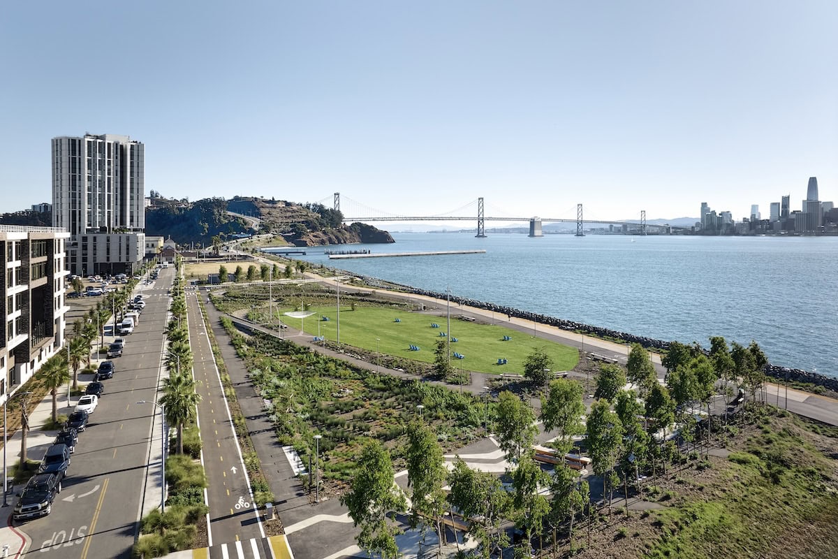 Aerial view of a waterfront park and residential buildings along the San Francisco Bay with the Bay Bridge and city skyline in the distance.