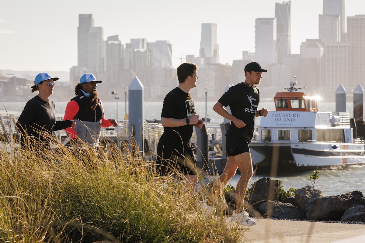 Four people jogging along a waterfront path with a city skyline and a docked boat in the background on a sunny day.