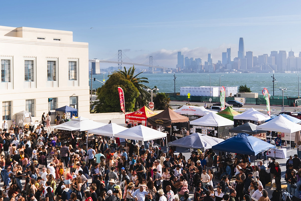 Crowd gathered at an outdoor market and event at Gold Bar on Treasure Island, with vendor tents, bay views, the Bay Bridge, and the San Francisco skyline in the background.
